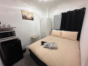 Une chambre d’hôtel de taille réduite, comprenant un lit, une armoire, une petite kitchenette et un tableau représentant un paysage urbain accroché au mur.