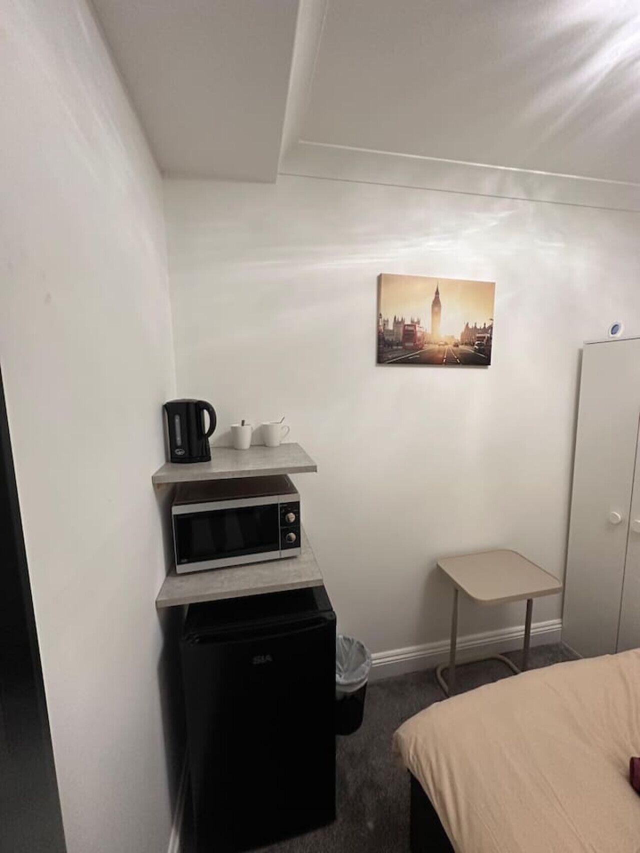 Quarto de hotel pequeno com cozinha compacta, uma cama, uma cadeira e um quadro com vista para a cidade na parede.