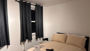Een slaapkamer met een bed, beige beddengoed, een radiator en zwarte gordijnen.