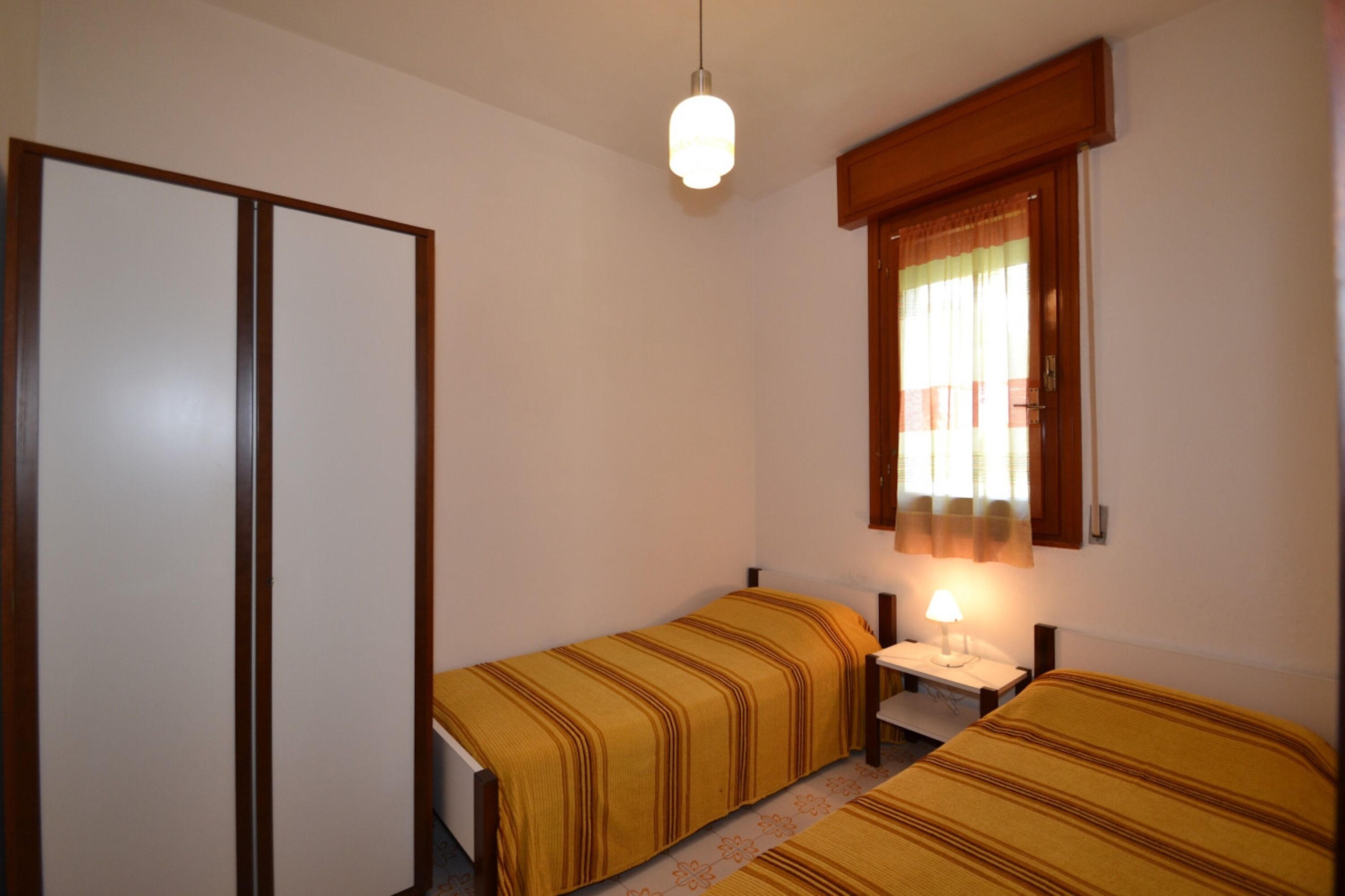 Ein Zimmer mit zwei Betten, einem Nachttisch, einer Lampe, einem Fenster mit Vorhängen und einer Pendelleuchte.