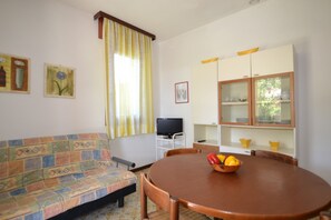 A room with a sofa, a dining table with chairs, a television on a stand, and a cabinet with glass doors. - Cozy Villa With Garden (San Michele al Tagliamento)