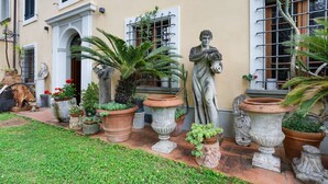 Ein Innenhof mit Kübelpflanzen, einer Statue und einer Gebäudefassade mit Fenstern und Fensterläden.
