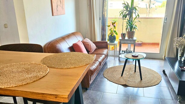 Een woonkamer met een bruine leren bank, een houten eettafel met geweven placematen en een glazen salontafel. Er is een schuifpui die leidt naar een balkon met potplanten.