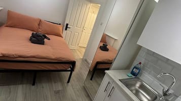 A compact room with a bed, a small kitchenette, and a door leading to another room.
