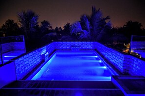 Ein rechteckiger Pool mit blauer Beleuchtung, umgeben von Steinmauern und Palmen.