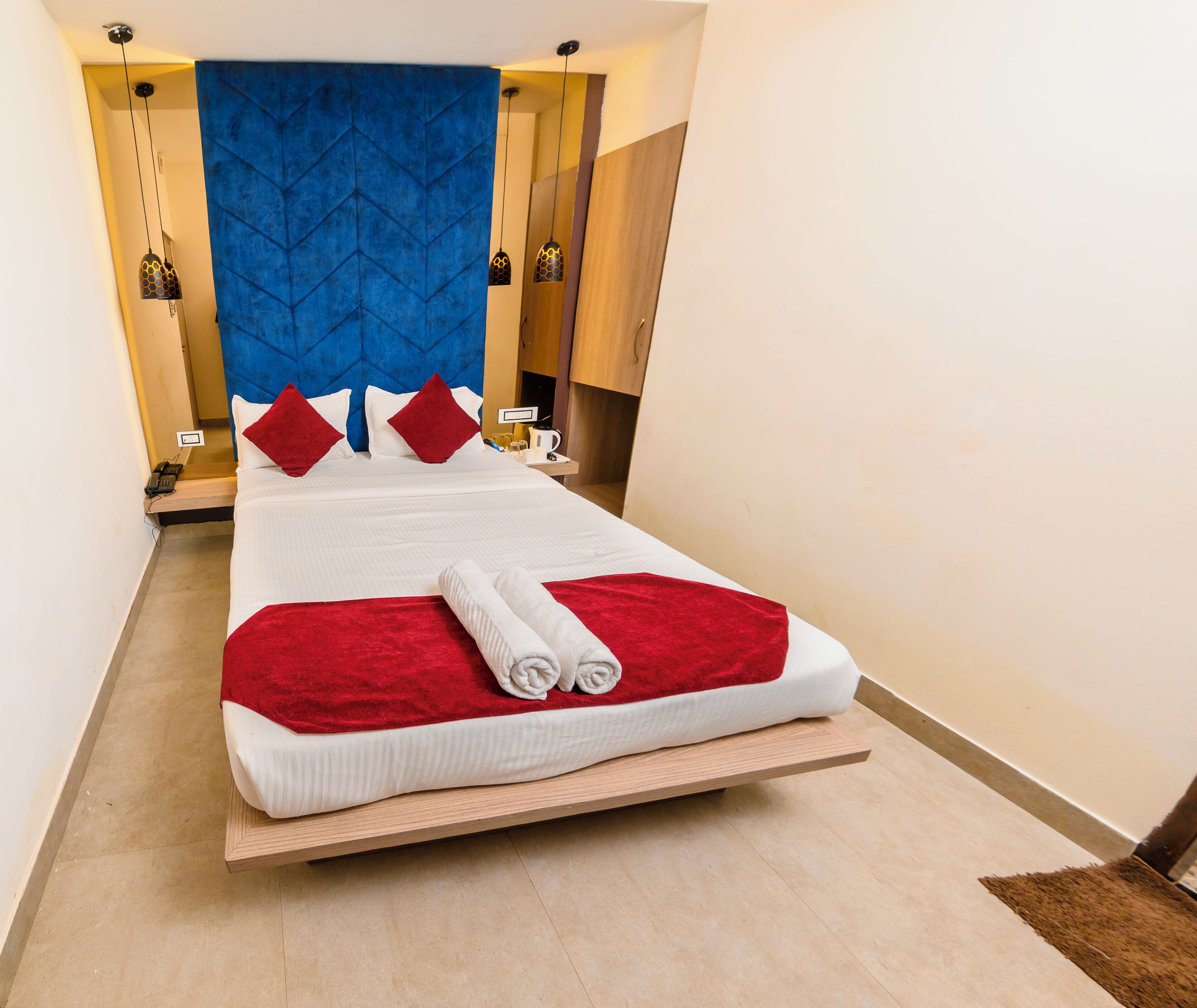 Un lit bien fait, recouvert d’une couvre-lit rouge et blanche, avec une tête de lit bleue et une table de chevet en bois sur laquelle se trouvent une lampe et une tasse.