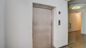 Pasillo con puerta cerrada del ascensor, un detector de incendios y un letrero que indica la dirección al ascensor.