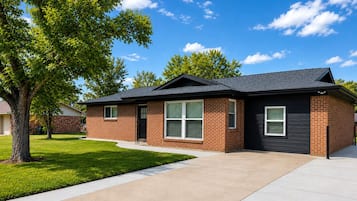 A two-story house with a brick exterior, a black roof, and a front yard with a tree and grass.