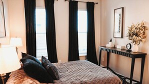Un dormitorio con una cama grande, cortinas oscuras, una mesita de noche con una lámpara, una pequeña estatua y un jarrón con plantas secas.