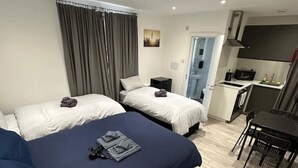 Ein Hotelzimmer mit zwei Betten, einem Schreibtisch, einem Stuhl, einer kleinen Küchenzeile und einer Tür zu einem weiteren Raum.