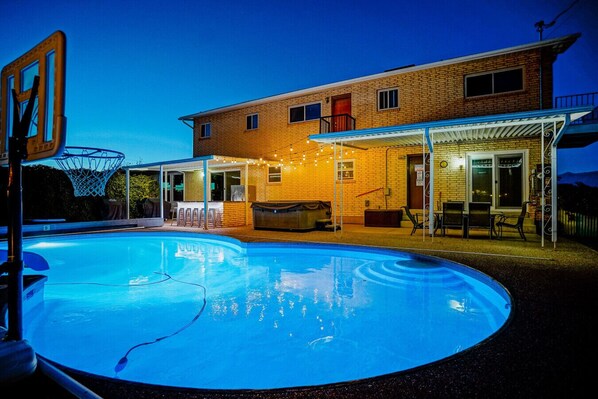 Una piscina con aro de baloncesto, un patio cubierto con luces colgantes y una casa de ladrillo al fondo.