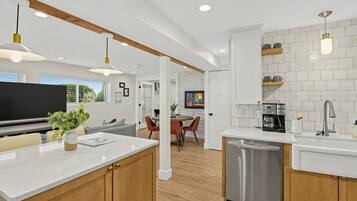 A modern kitchen with white countertops, wooden cabinets, and stainless steel appliances. There is a dining area with a table and chairs, and a television mounted on the wall.