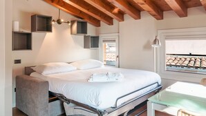 A modern bedroom with a bed, bedside tables, a glass table, and a window with a view of the roof. - Suite Cavour (Brescia)