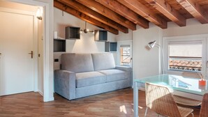 A modern living room with a glass table, wicker chairs, a grey sofa, wooden flooring, and exposed wooden beams. - Suite Cavour (Brescia)