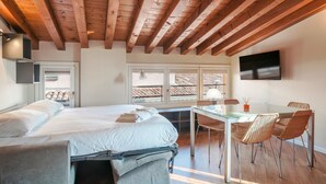 A bedroom with a bed, a desk, and chairs, wooden beams on the ceiling, a TV on the wall, and a window with a view of the outside. - Suite Cavour (Brescia)