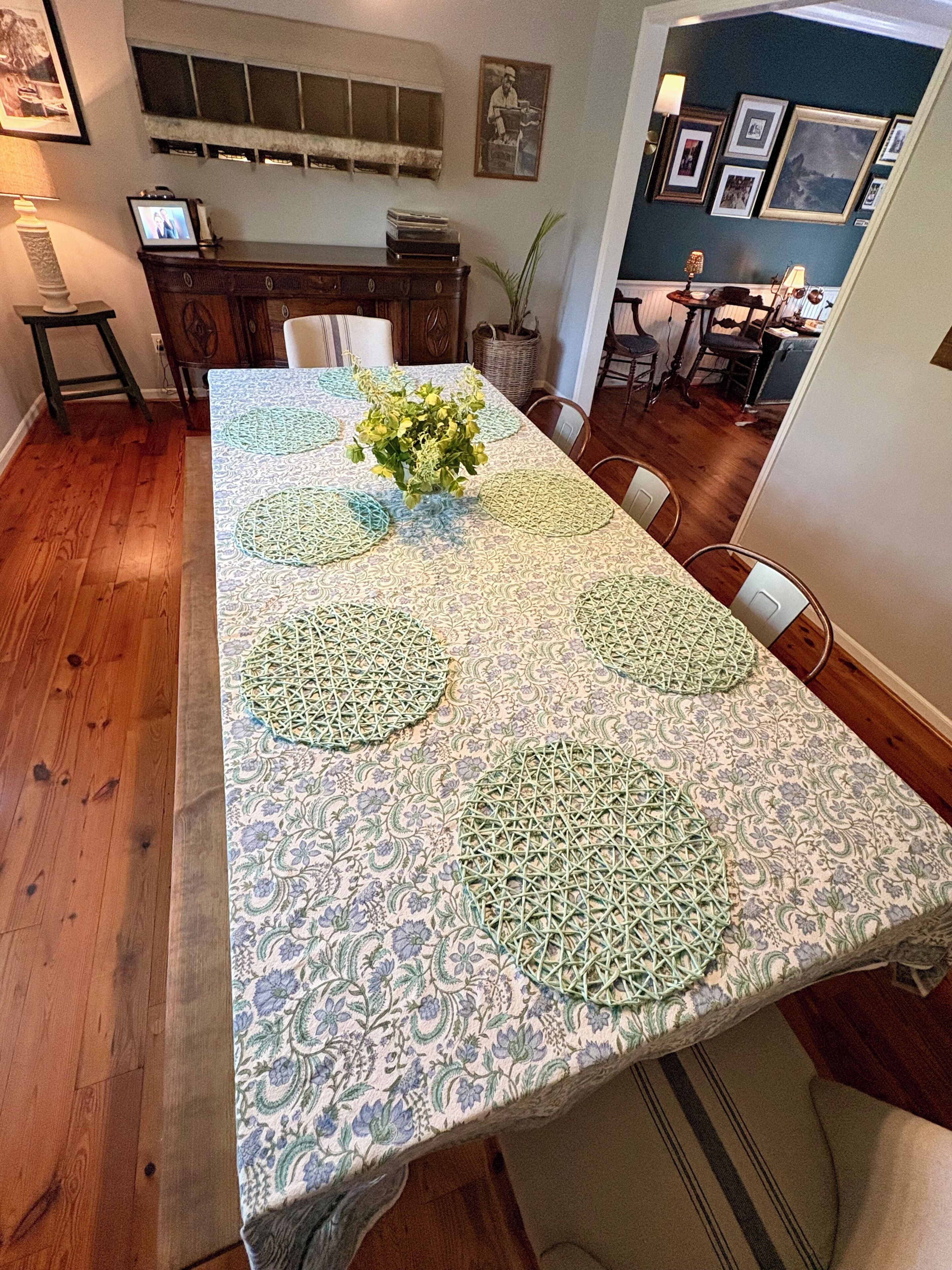A dining table with a patterned tablecloth, surrounded by chairs, in a room with wooden flooring and a cabinet.