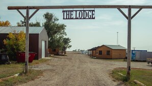 Un camino de grava que conduce a un edificio rústico con el rótulo 'THE LODGE', techo de metal, estructuras de madera y un contenedor de basura rojo.