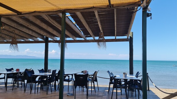 Ein Sitzbereich im Freien mit Tischen und Stühlen unter einer Holzw pergola, mit Blick auf ein ruhiges Meer und entfernte Berge.