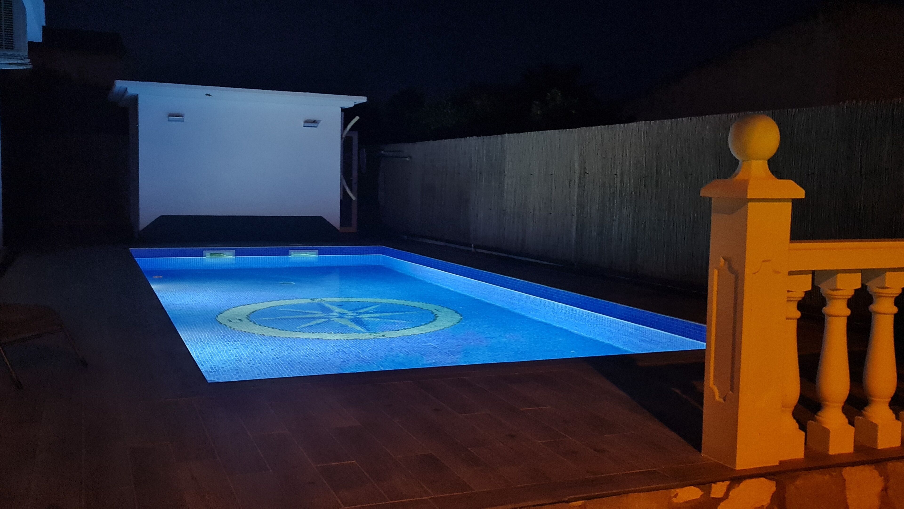 A rectangular swimming pool with a circular design in the center, surrounded by a wooden deck and a concrete wall.