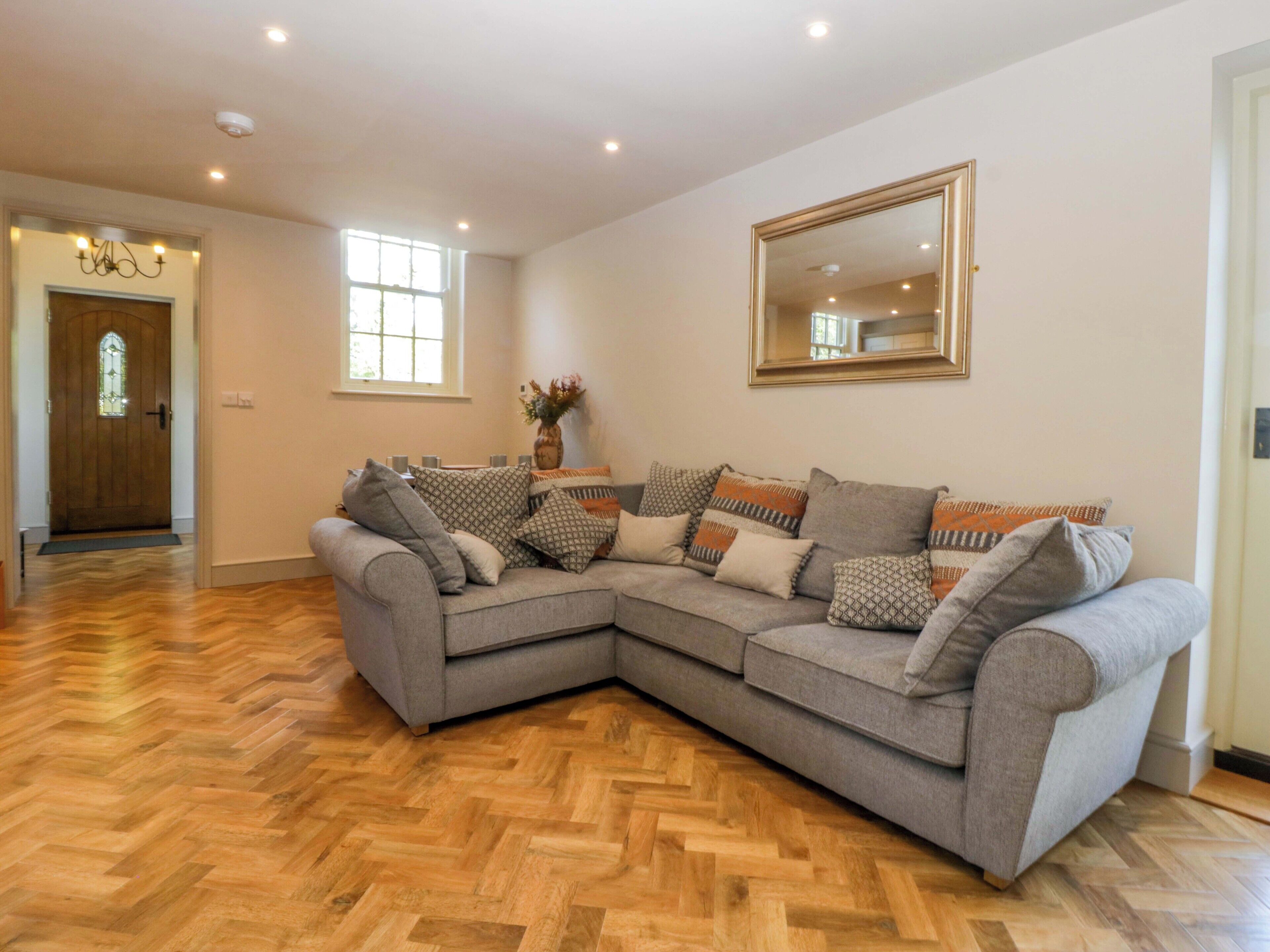 A spacious living room with a large L-shaped grey sofa adorned with patterned cushions, a wooden herringbone floor, a large mirror on the wall, and a door leading to another room.