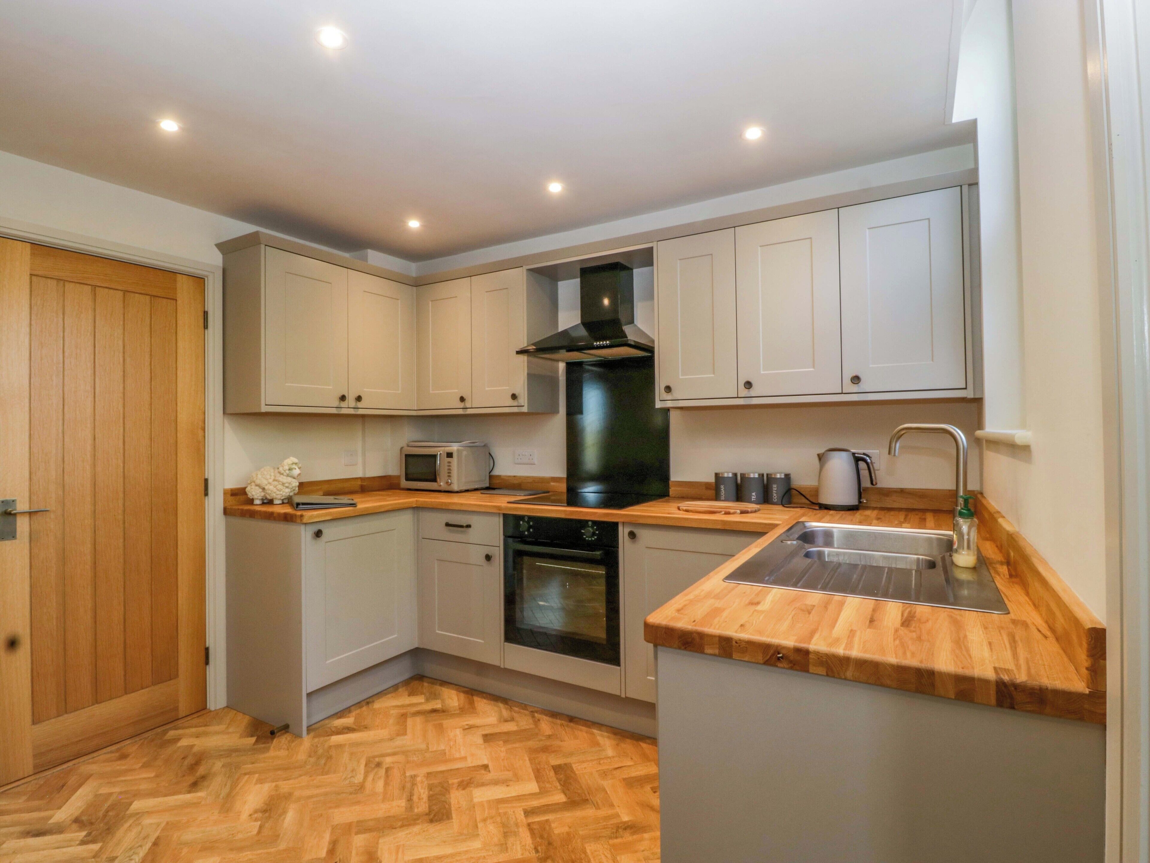 Una cocina compacta con suelo de madera, armarios blancos, horno empotrado, microondas y un fregadero.