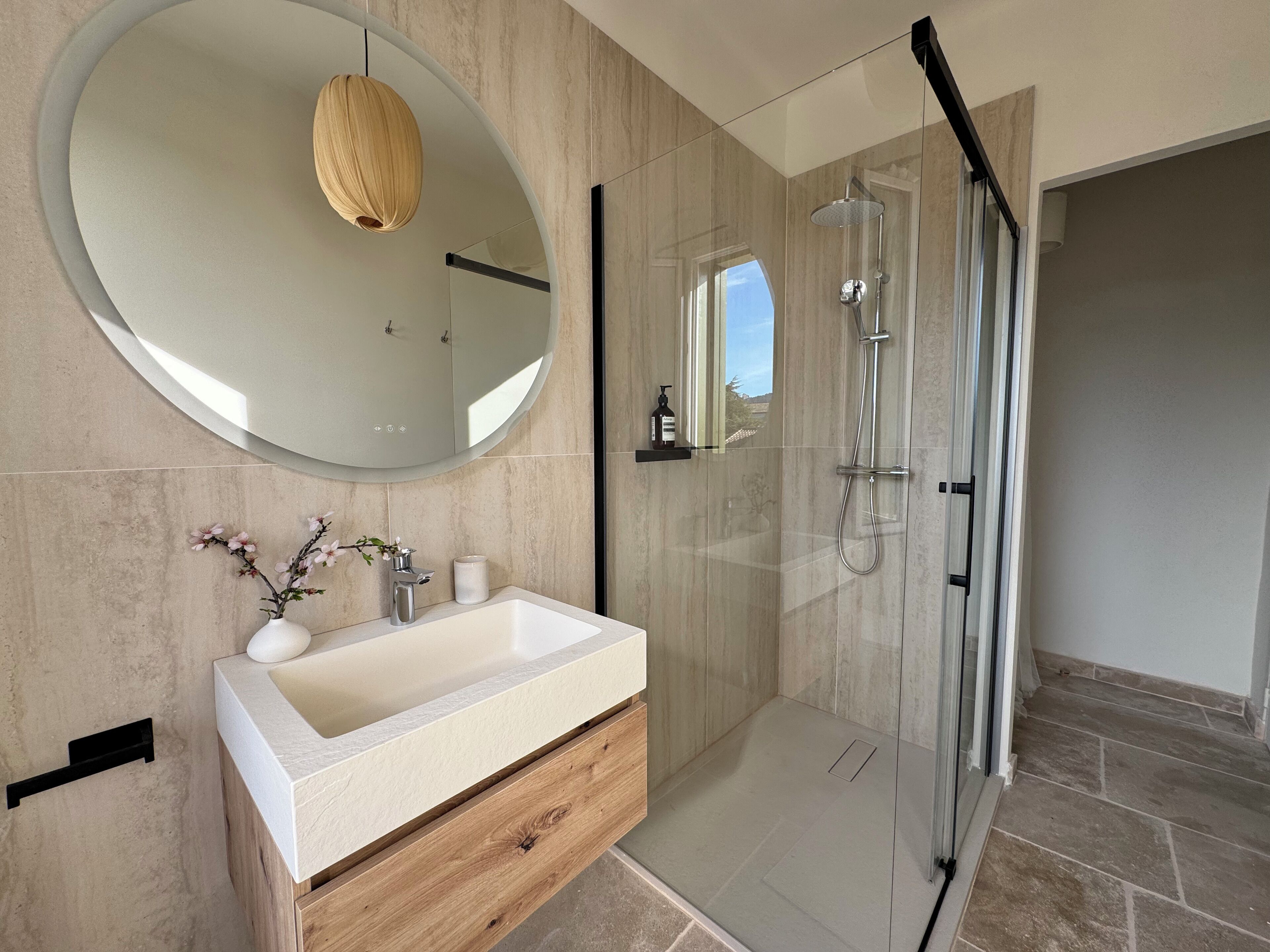 Een moderne badkamer met een grote ovale spiegel, een houten wastafelmeubel, een douchecabine van glas en uitzicht op de omgeving.