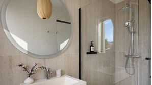 Um banheiro moderno com um espelho redondo grande, uma pia com um vaso de flores, um chuveiro com chuveiro manual e uma garrafa de sabonete em uma prateleira.
