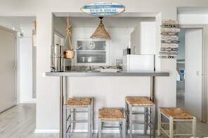 A compact kitchen with a hanging light fixture, a stainless steel kettle, and a wooden bowl on the counter. There are three stools with woven seats. - BLAUMAR 7G Roses -Immo Barneda (Rosas / Roses)