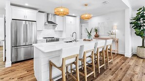 A modern kitchen with white cabinetry, stainless steel appliances, and a central island with bar stools. - Coastal Calm (Santa Rosa Beach)