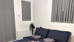 Ein Schlafzimmer mit Bett, Nachttisch, Ventilator und Fenster mit Jalousien.