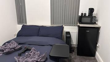 A neatly made bed with a dark blue comforter and pillows, a small black refrigerator with a coffee maker on top, and a compact black heater beside the bed.