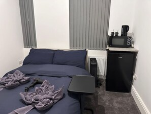 A neatly made bed with a dark blue comforter and pillows, a small black refrigerator with a coffee maker on top, and a compact black heater beside the bed. - Buckingham Palace/Hyde Park near (Greater London)
