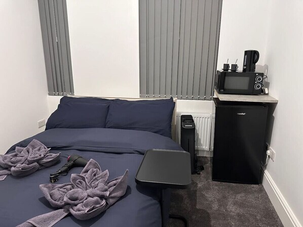 A neatly made bed with a dark blue comforter and pillows, a small black refrigerator with a coffee maker on top, and a compact black heater beside the bed. - Buckingham Palace/Hyde Park near (Greater London)