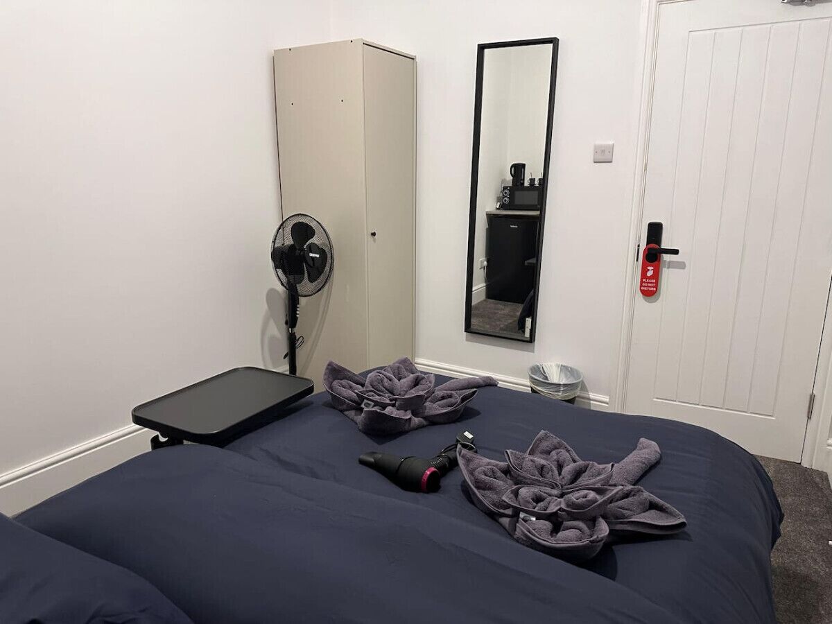 A hotel room with a bed, a bedside table, a fan, a mirror, a wardrobe, and a door with a red emergency exit sign.