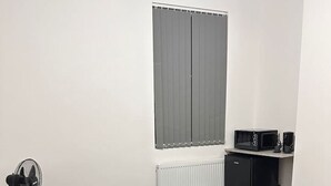 Een slaapkamer met een bed, een radiator, een raam met gordijnen, een kleine tafel en een magnetron.