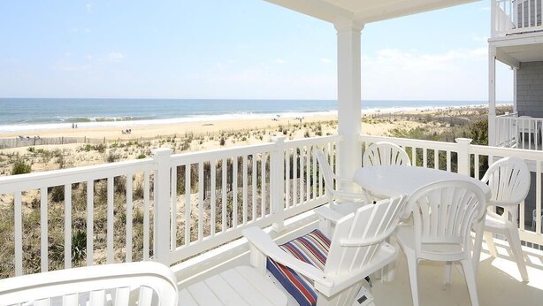 A covered balcony with white chairs and a table, overlooking a beach with sand dunes and the ocean. - Oceanfront 3BR Condo in Fenwick | Walk to Beach! (Fenwick Island)