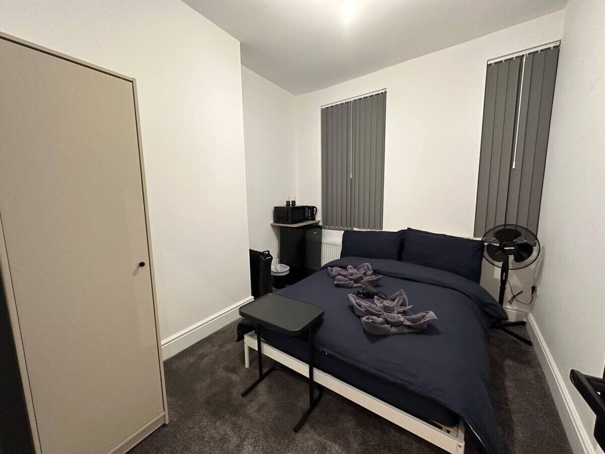 A bedroom with a bed, a desk, a chair, a radiator, and a window with blinds.