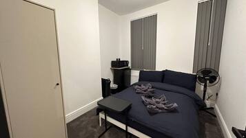 A bedroom with a bed, a desk, a chair, a radiator, and a window with blinds.
