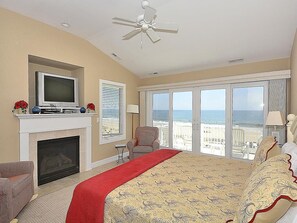 Ein Schlafzimmer mit Kamin, Fernseher, Bett mit rotem Läufer, Sessel und Blick auf den Strand.