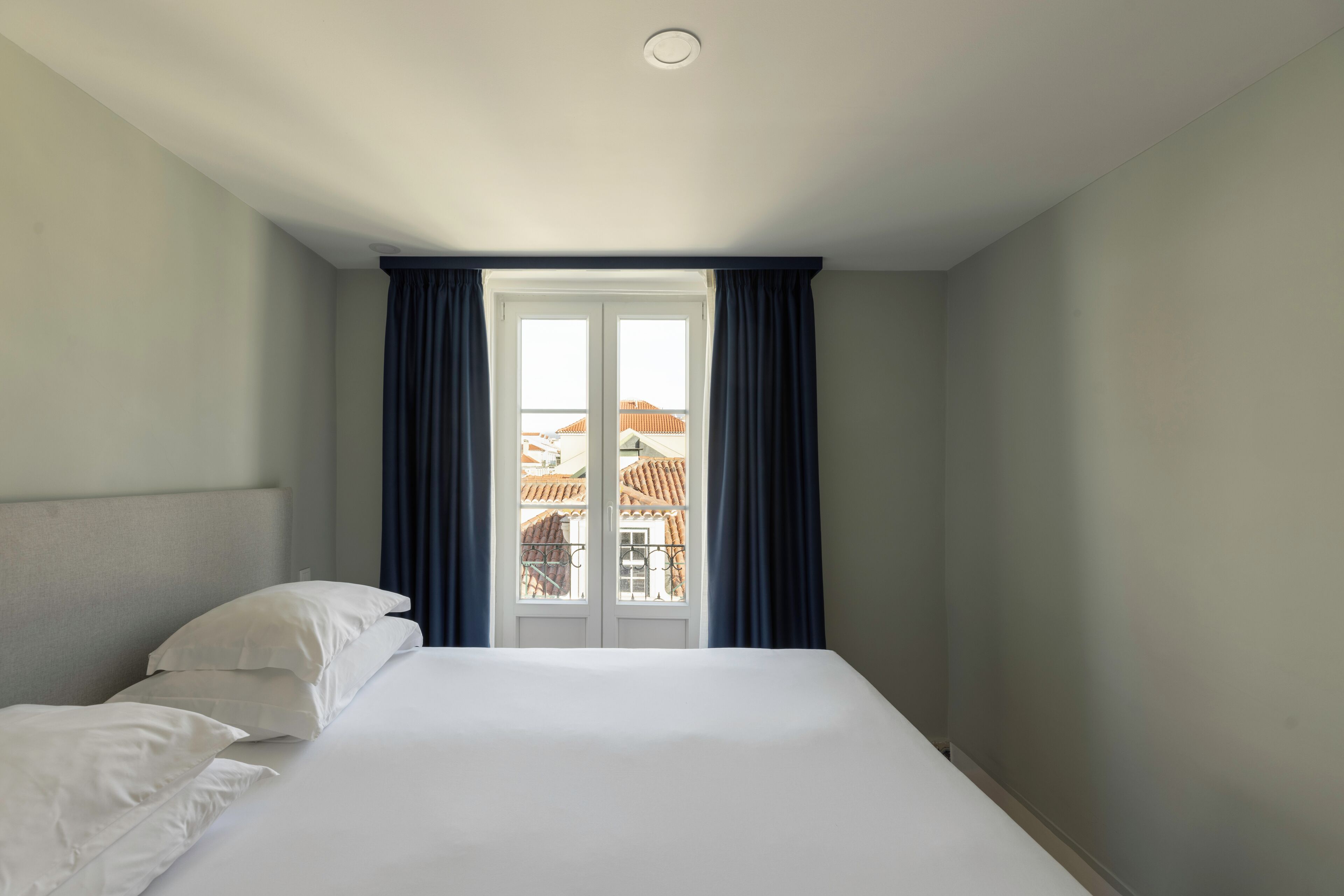 Une chambre d’hôtel avec un lit, des oreillers blancs, une tête de lit, une fenêtre avec des rideaux et une vue sur les bâtiments environnants.