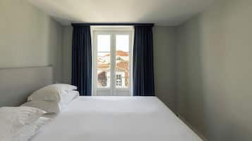 Une chambre d’hôtel avec un lit, des oreillers blancs, une tête de lit, une fenêtre avec des rideaux et une vue sur les bâtiments environnants.
