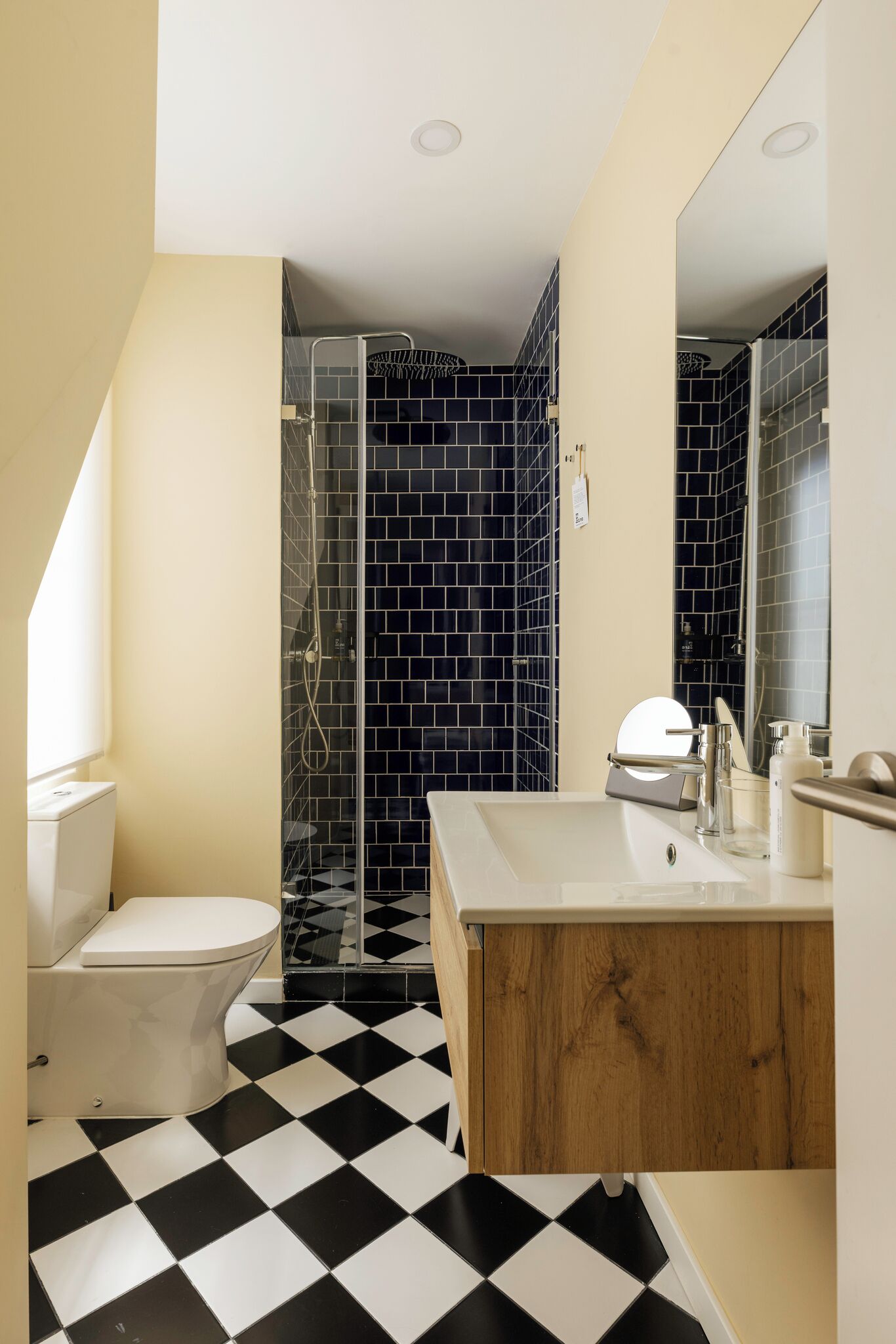 A bathroom with a black and white checkered floor, a white toilet, a wooden vanity with a mirror, and a glass shower enclosure.