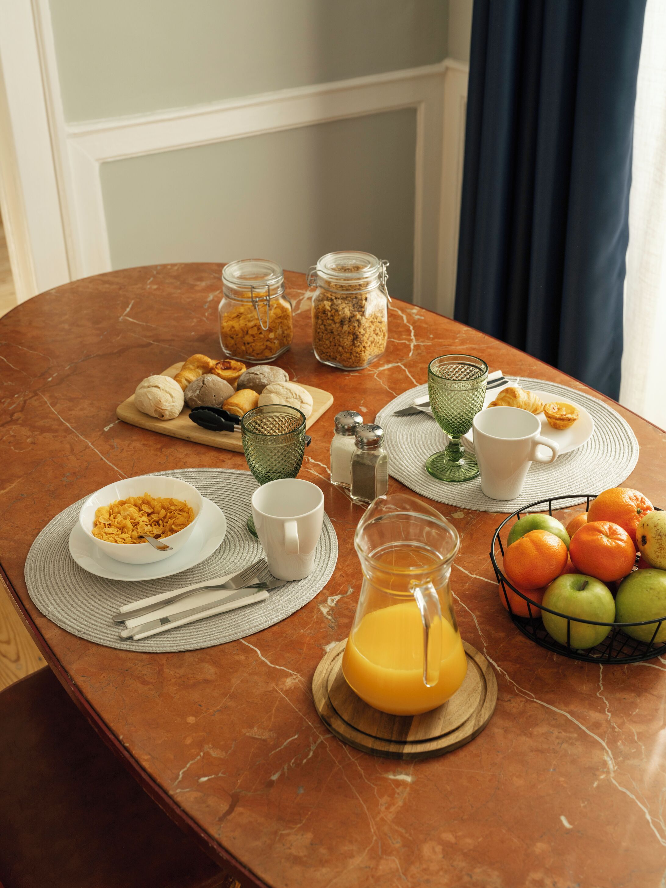 Una mesa de desayuno con diferentes alimentos, una jarra de jugo de naranja y una cesta de frutas.