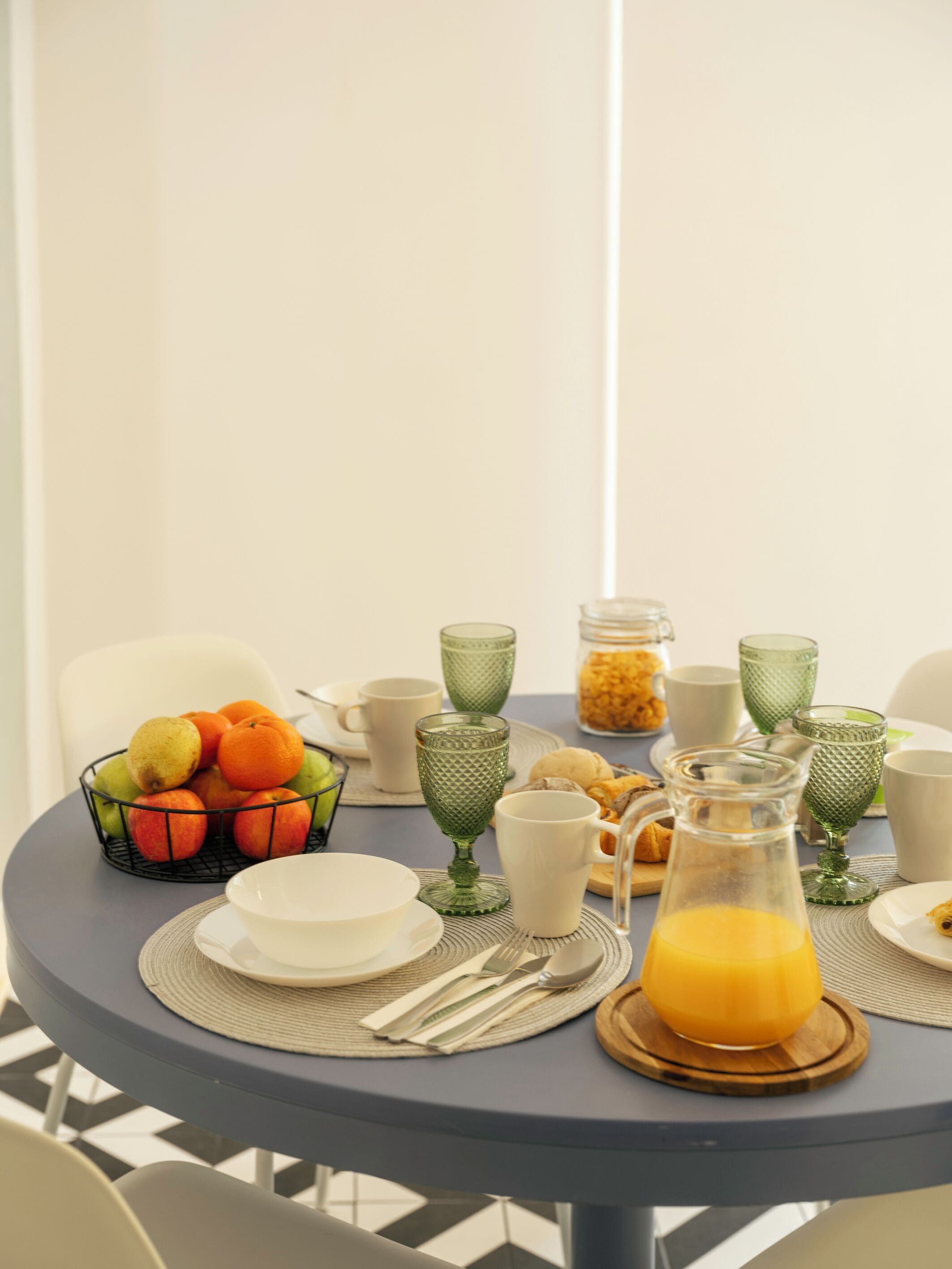 Una mesa de desayuno con un frutero, vasos de vidrio verde, platos blancos y una jofaina de jugo de naranja.