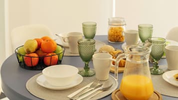 Una mesa de desayuno con un frutero, vasos de vidrio verde, platos blancos y una jofaina de jugo de naranja.