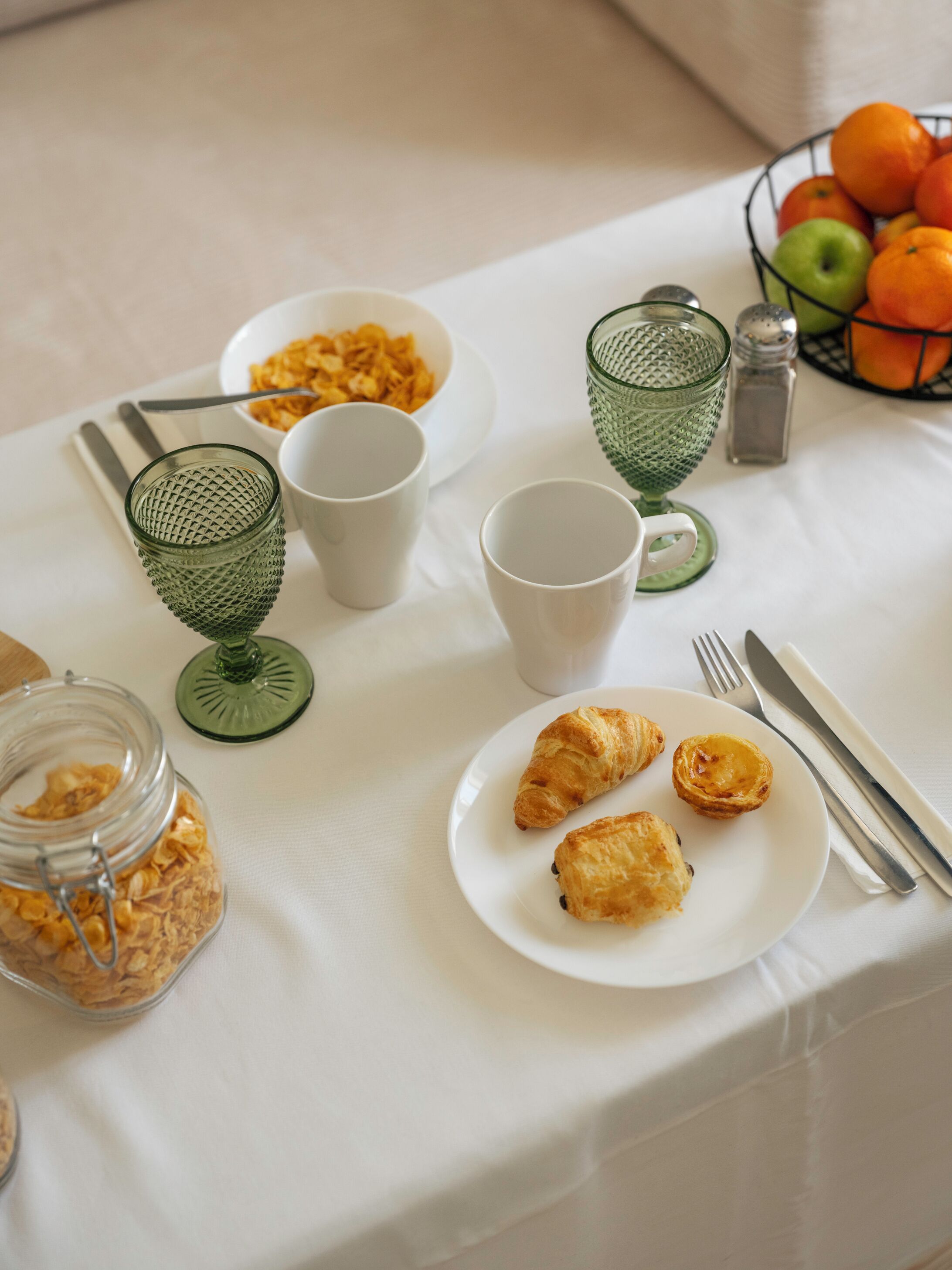 A breakfast setting with a plate of pastries, a bowl of cereal, and a glass jar of cereal flakes.