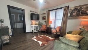 Una sala de estar con piso de madera, un sofá verde, un tapete de cuero de vaca, un televisor, una mesita y una ventana con cortinas.