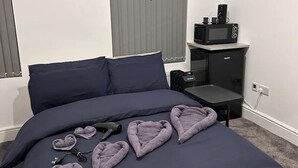 Una cama bien hecha con un edredón azul oscuro, una mesita de noche con microondas y almohadas con forma de corazón sobre la cama.
