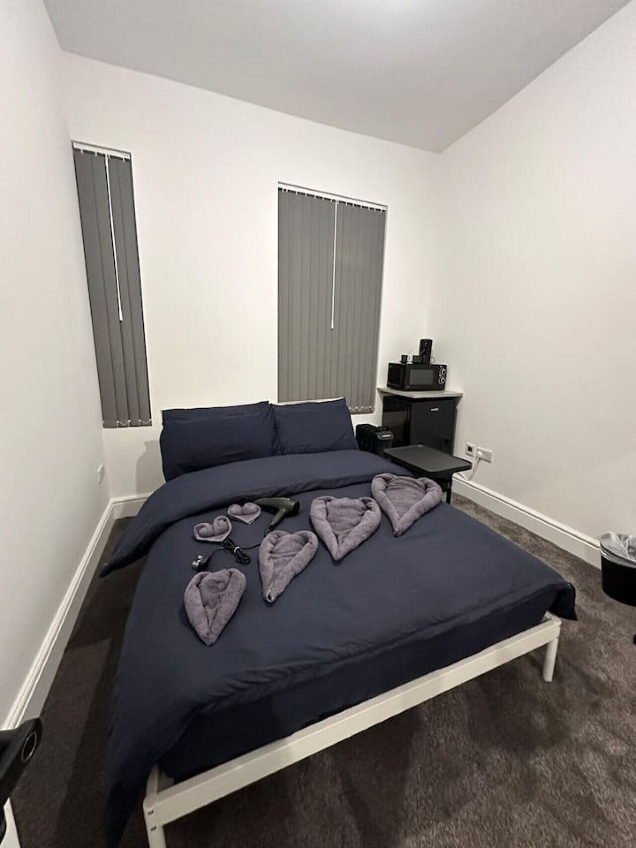 A bedroom with a bed, a bedside table, a TV, and two windows with blinds.