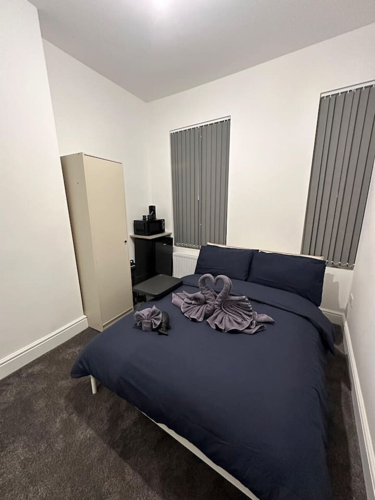 A hotel room with a bed, a wardrobe, a small table with a kettle, and a window with blinds.